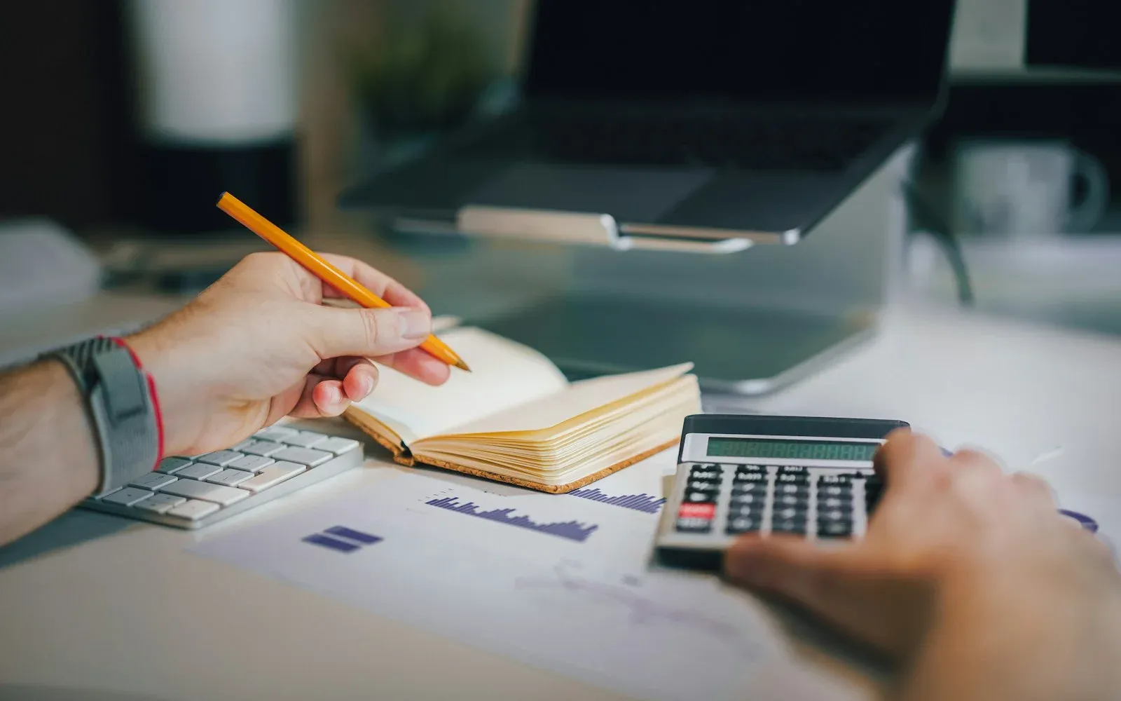 A person writing in a notebook while working a calculator, with a laptop and a chart printout on the desk
